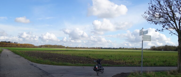 Landschaft mit S-Pedelec an einer Wergabelung eines Fahrradweges zwischen Wiesen und Feldern